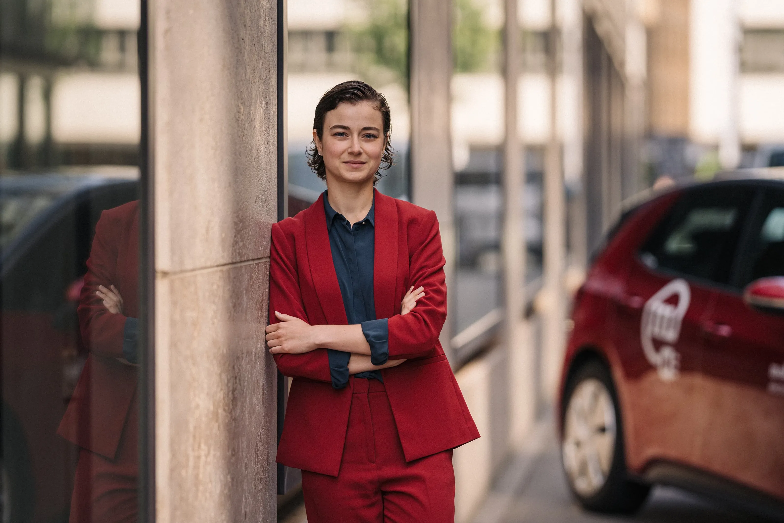 Woman in red suit leaning against the wall of the house next to a Mobility car sharing vehicle in the city.