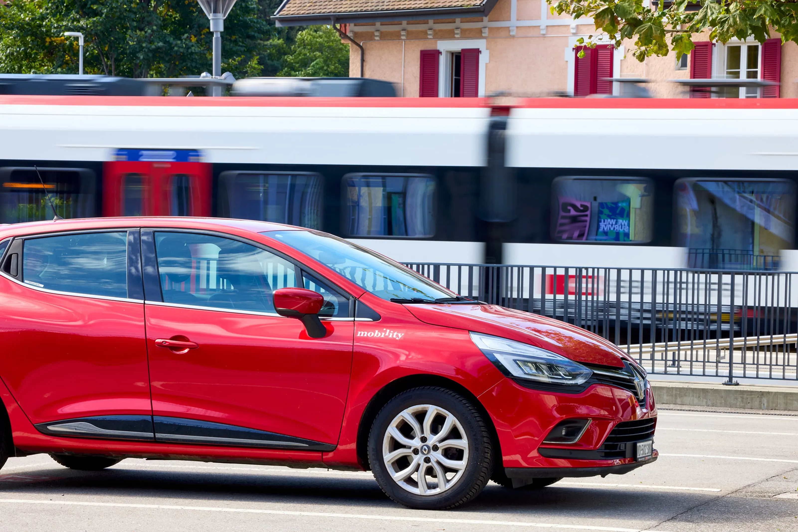 Red car sharing car driving along a road while an SBB S-Bahn train passes by in the background.