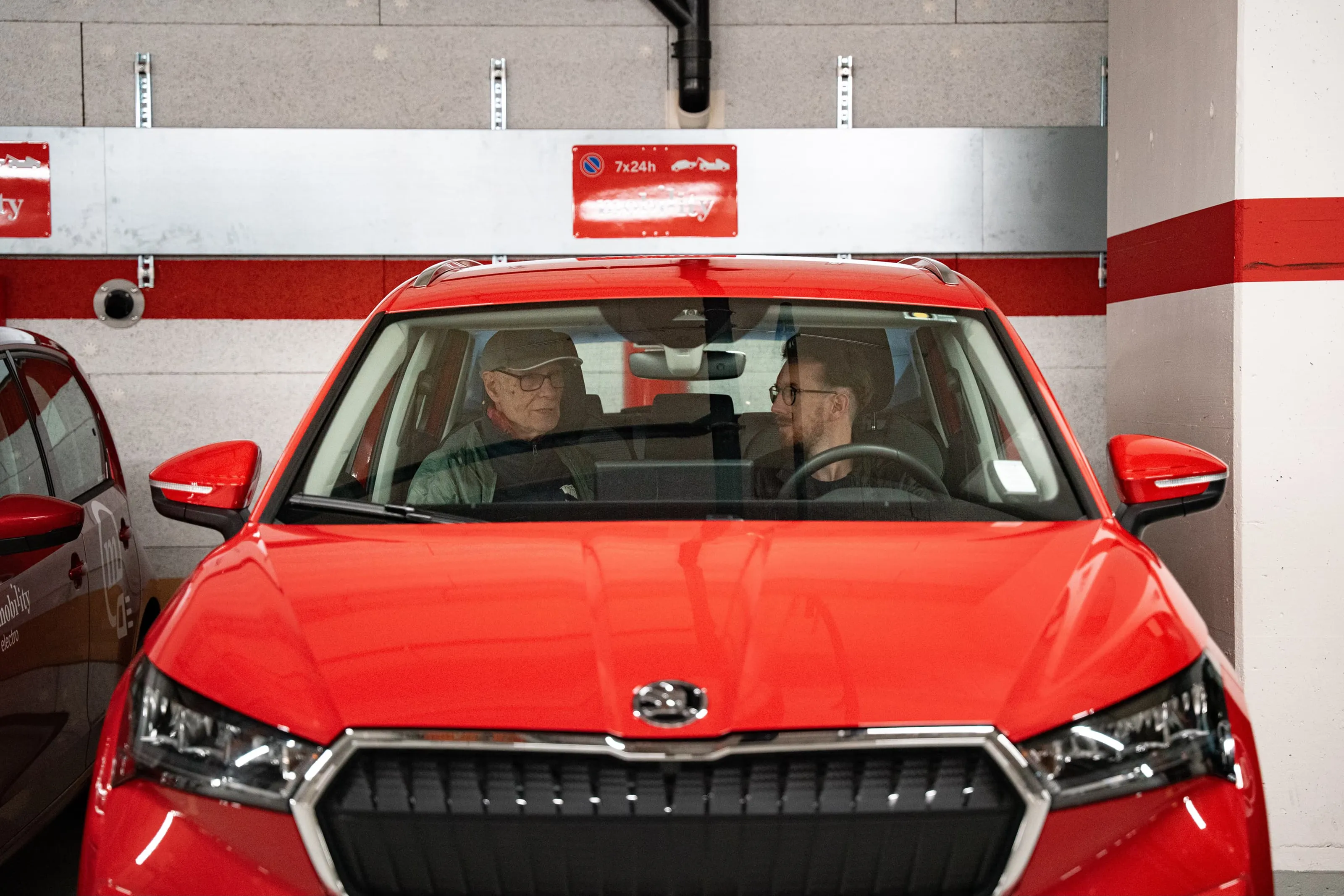 Two men talking while sitting in an underground car park in a red Mobility car.