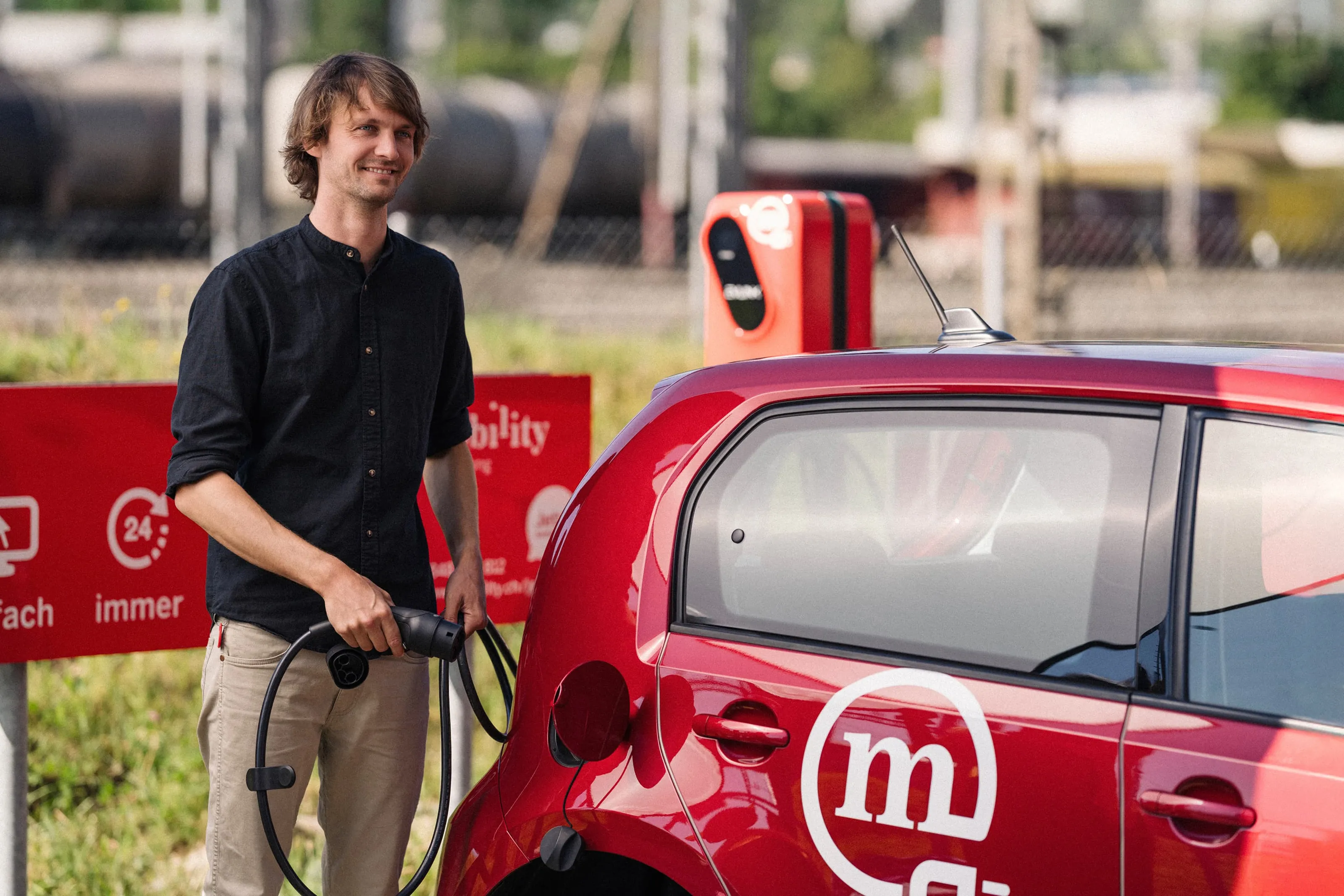 Man smiling while charging a red Mobility car sharing electric car at a public charging station.