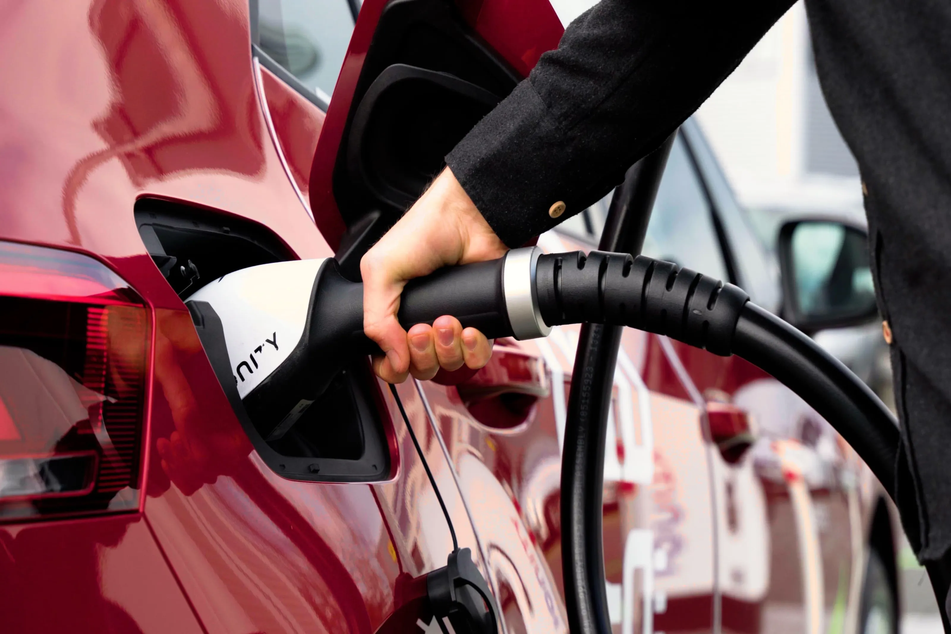 Hand plugs charging cable into a red electric car at a charging station.
