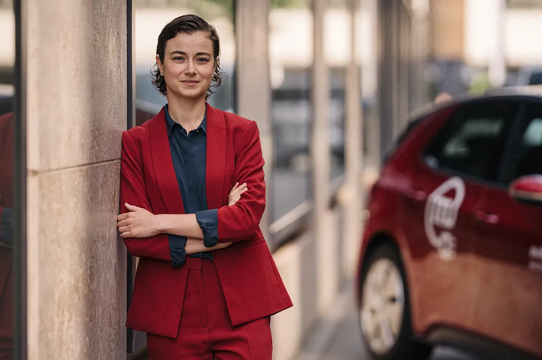 Woman in red suit leaning against the wall of the house next to a Mobility car-sharing vehicle in the city.