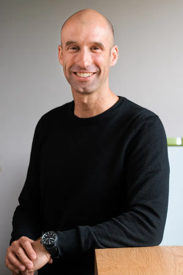 Laurent Roux in a black sweater smiling at the camera and leaning against a table in the office.