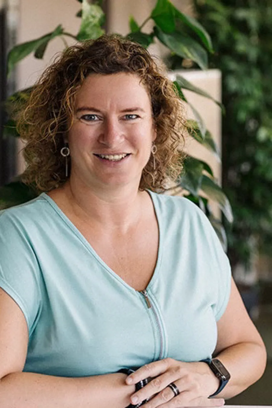 Woman with curly hair standing smiling in front of plants in an office.