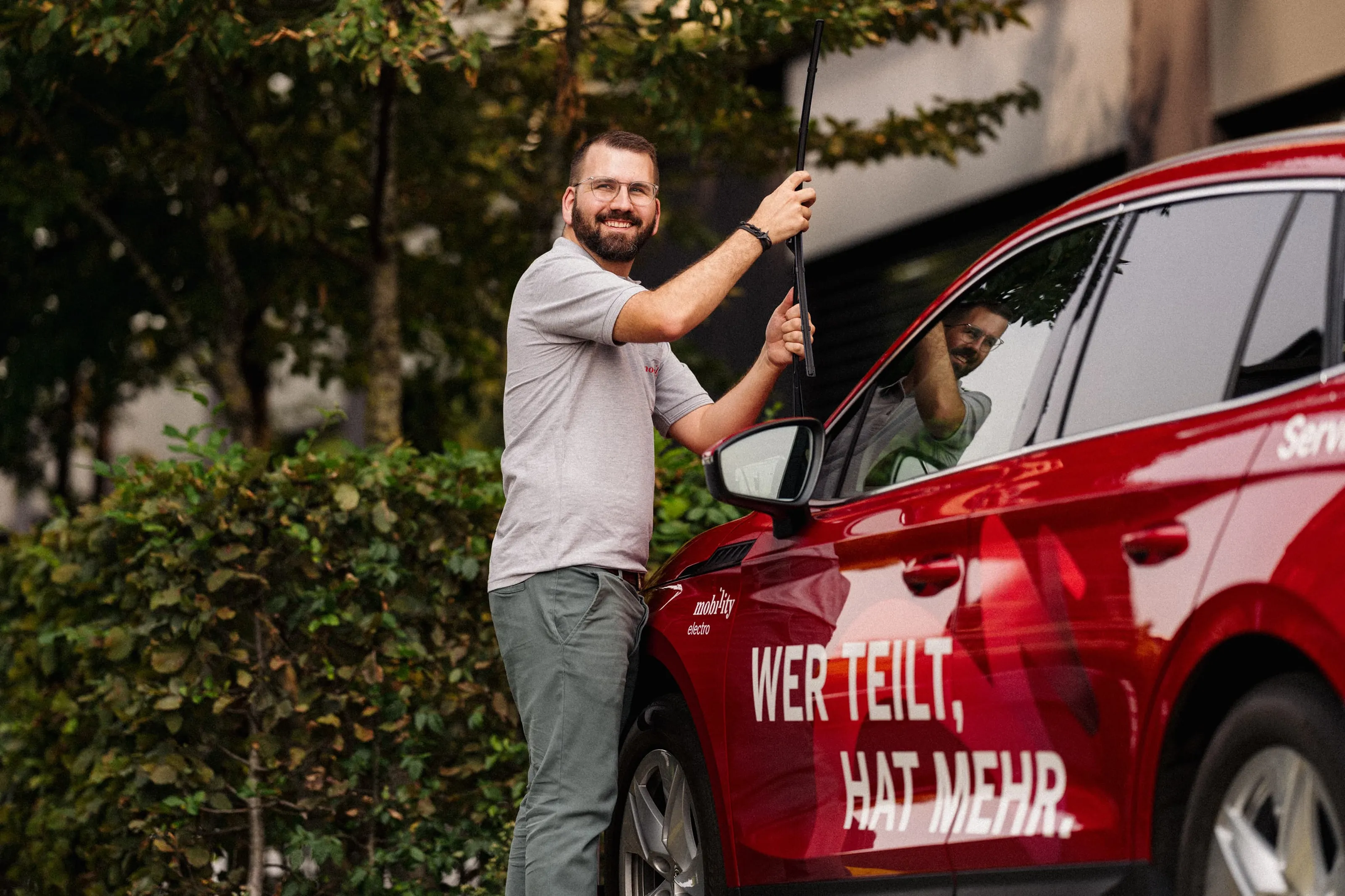 Man cleaning the windscreen wiper of a red Mobility car sharing car outdoors.