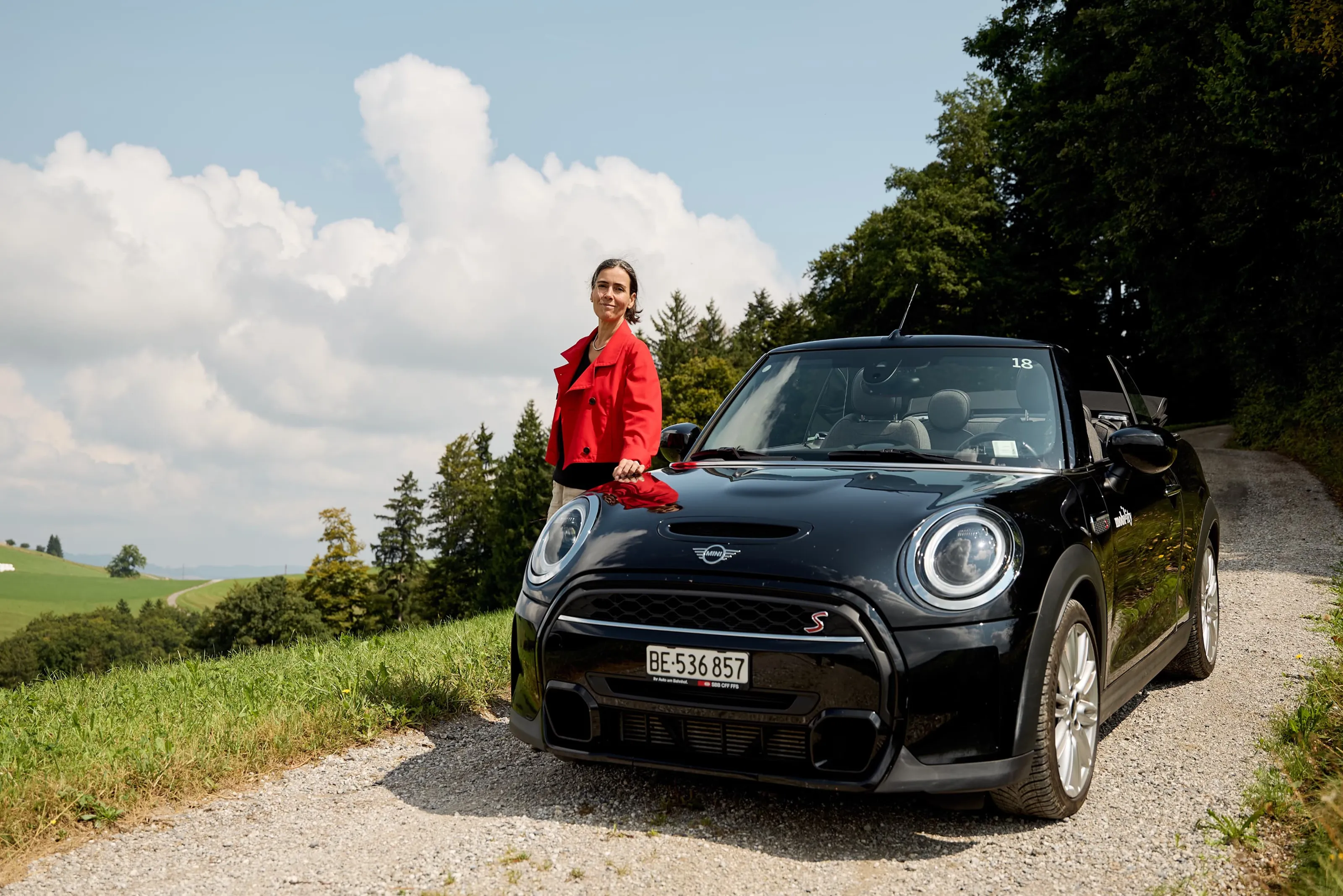 Woman standing next to a black convertible on a rural road overlooking meadows and trees.
