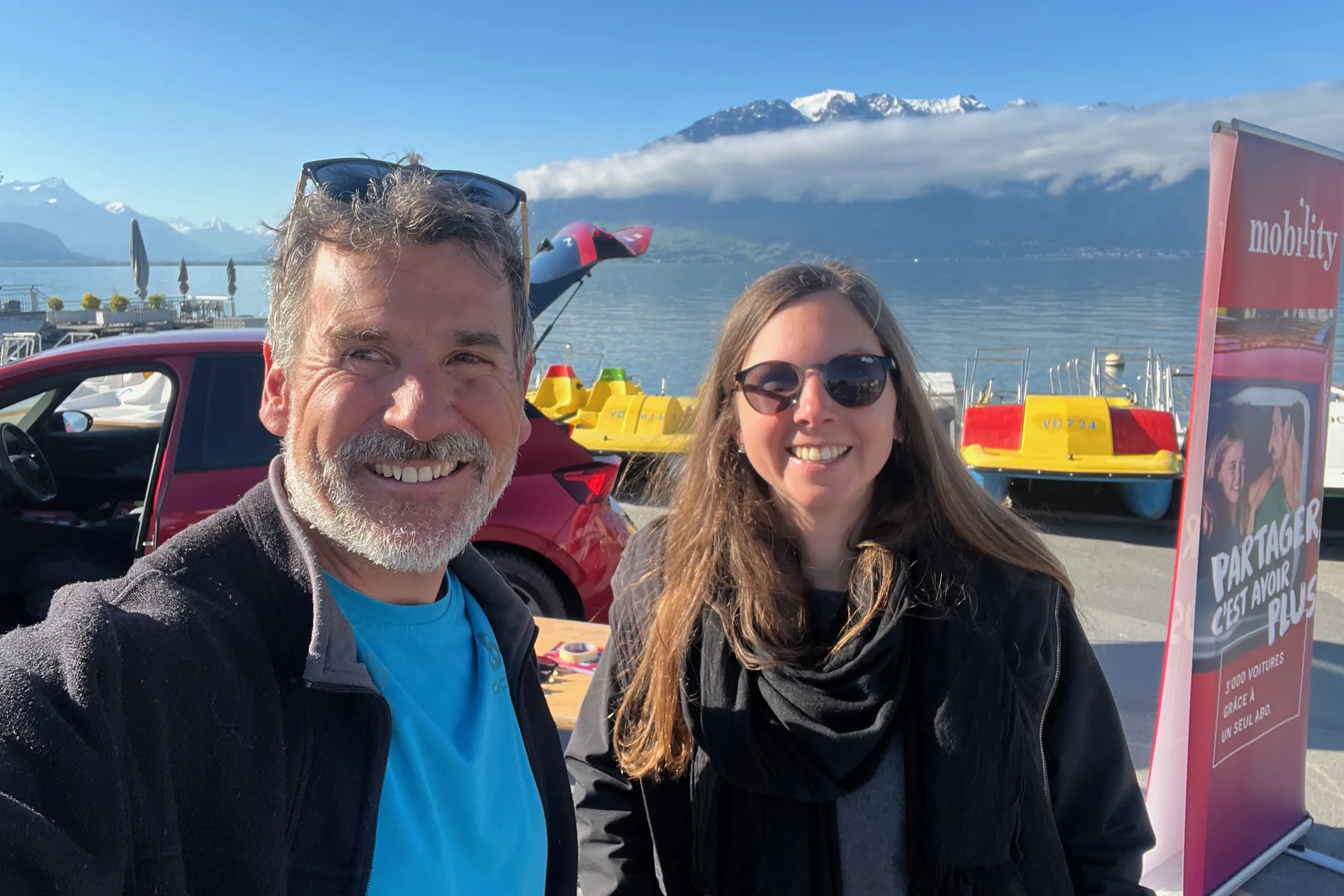 Two people taking a selfie by a lake in front of mountains next to a red car sharing car and pedal boats.