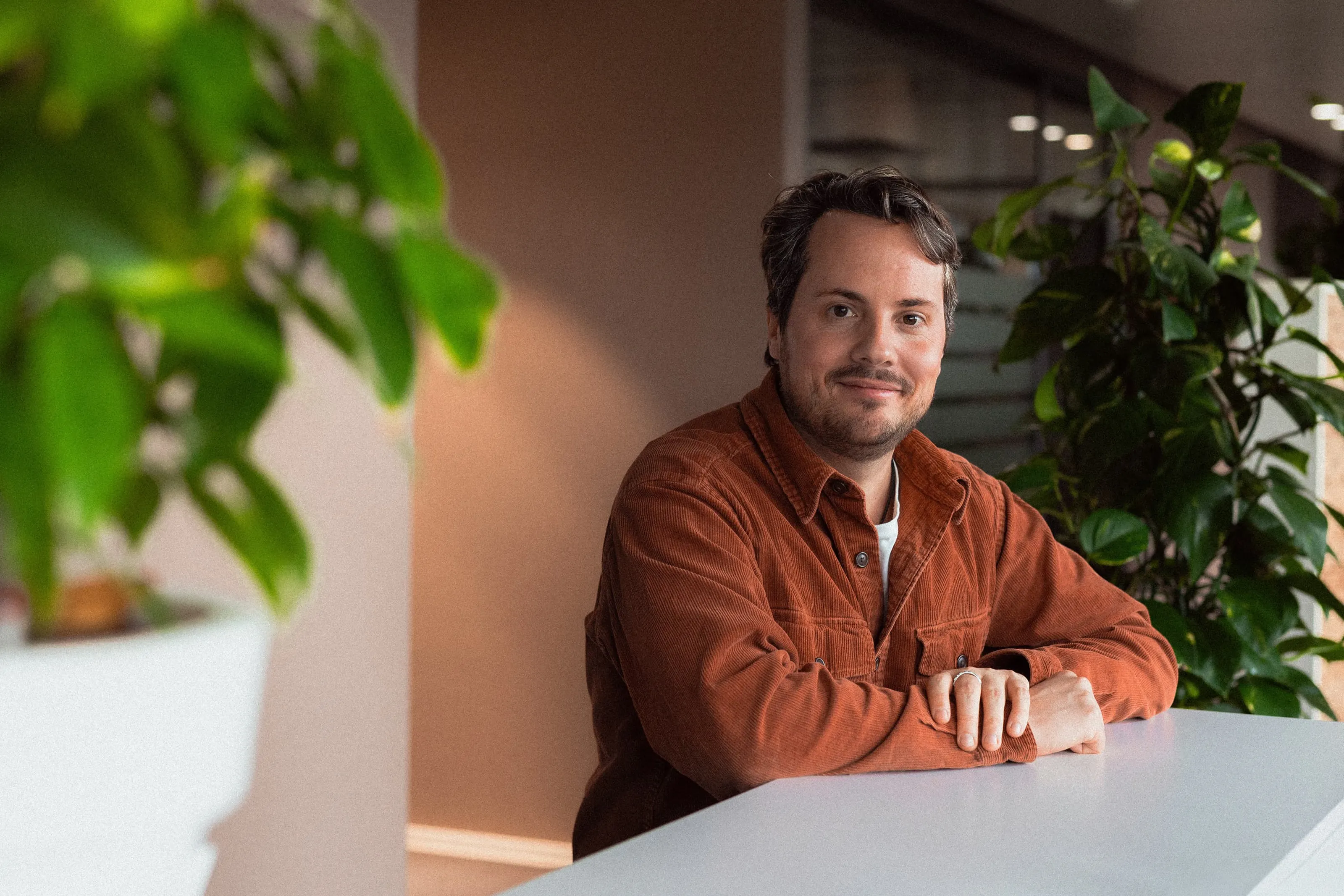 Man in a brown shirt sitting smiling at a table in a modern office with plants.