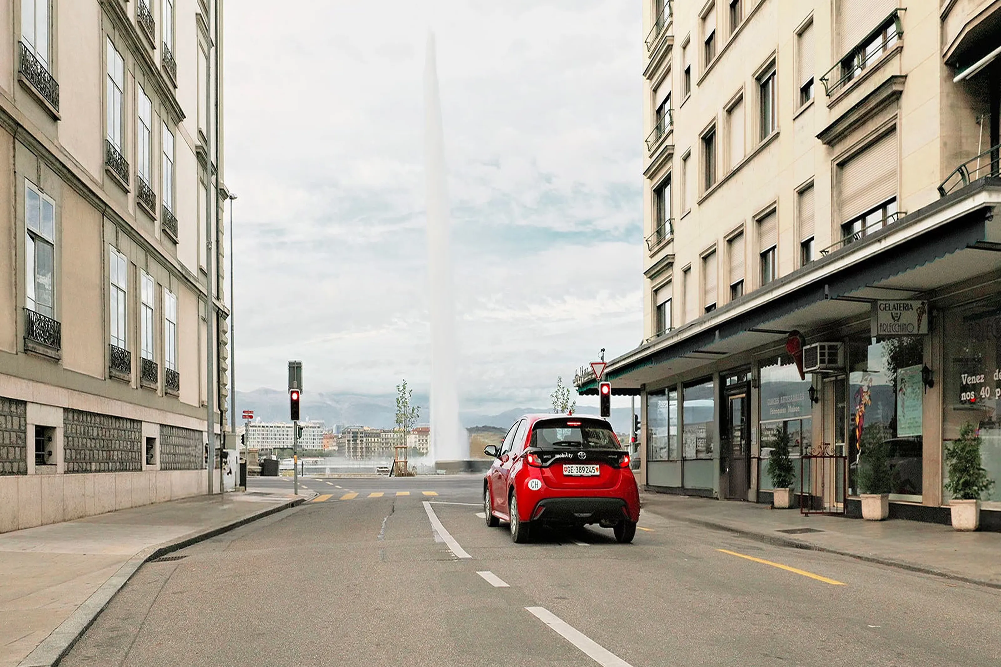 Red Mobility car sharing car driving along a city street towards the Jet d’Eau fountain in Geneva.